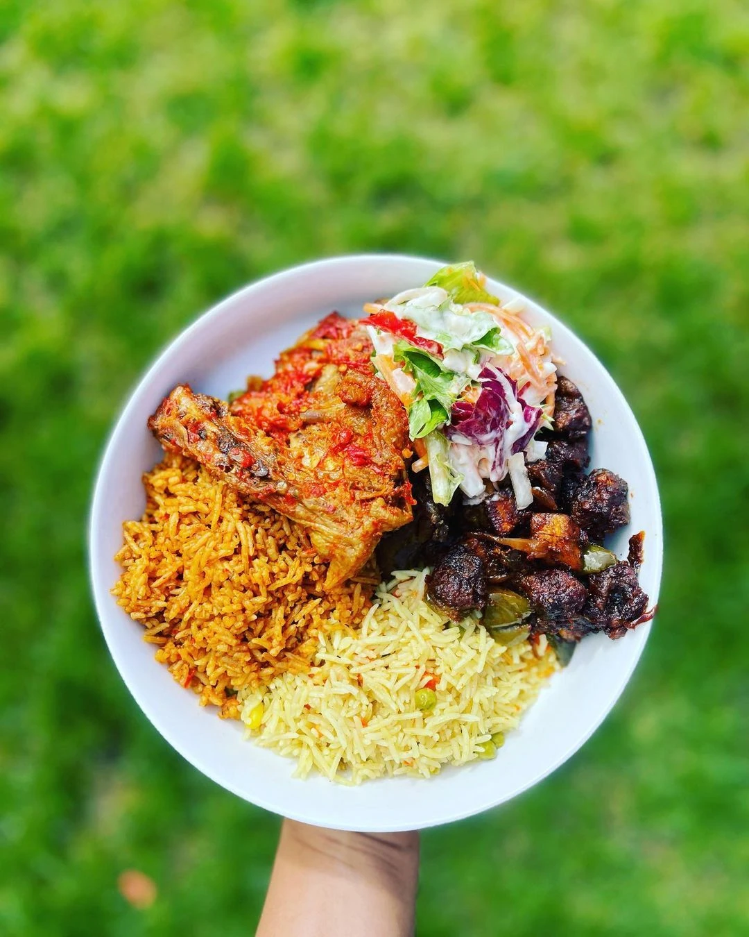 A white plate of jollof rice, fried rice, peppered chicken, plantain and salad